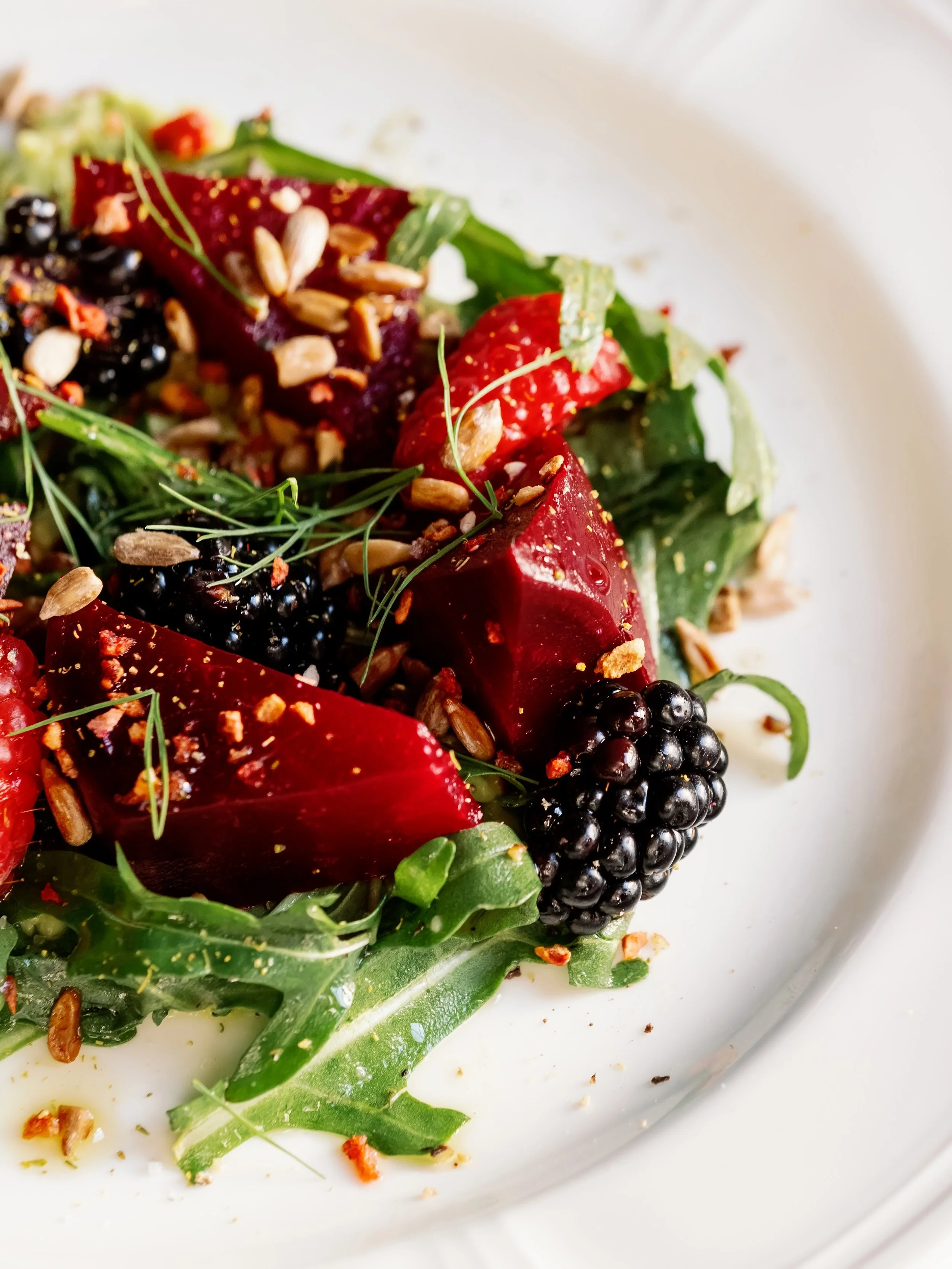 Close-up of a colorful salad with blackberries, beets, greens, sunflower seeds, and herbs on a white plate.
