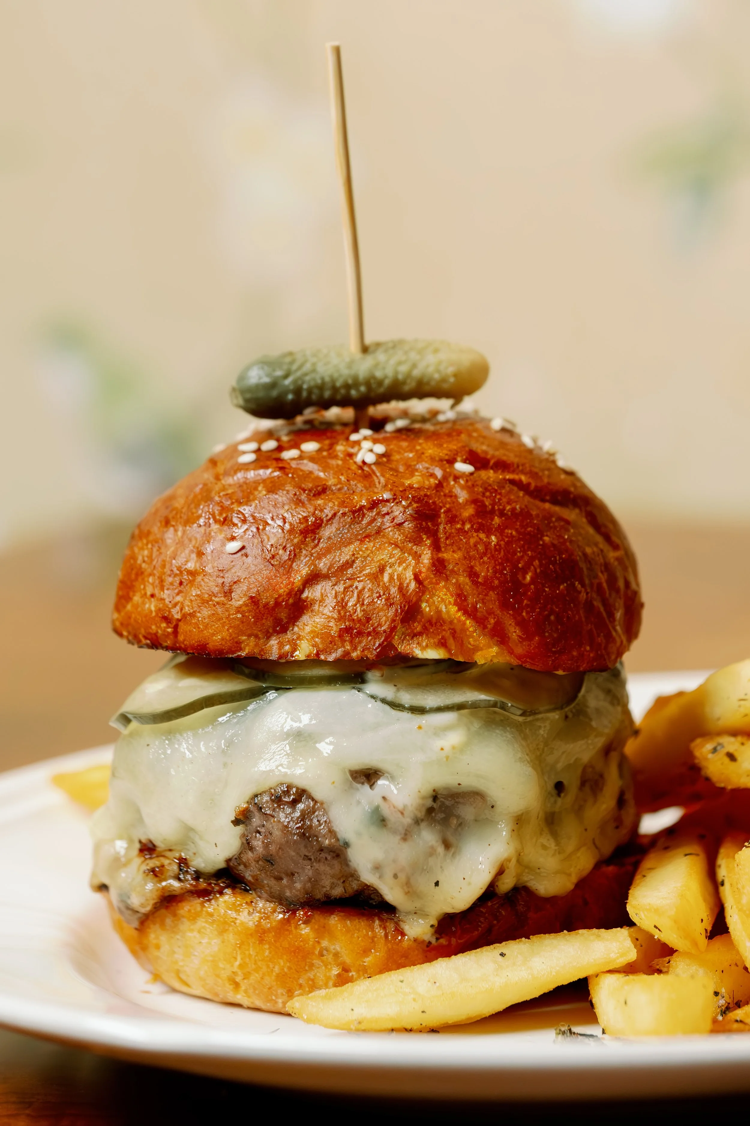 Close-up of a cheeseburger with melted cheese, pickles, and a sesame seed bun topped with an olive and a skewer, served with French fries on a white plate.