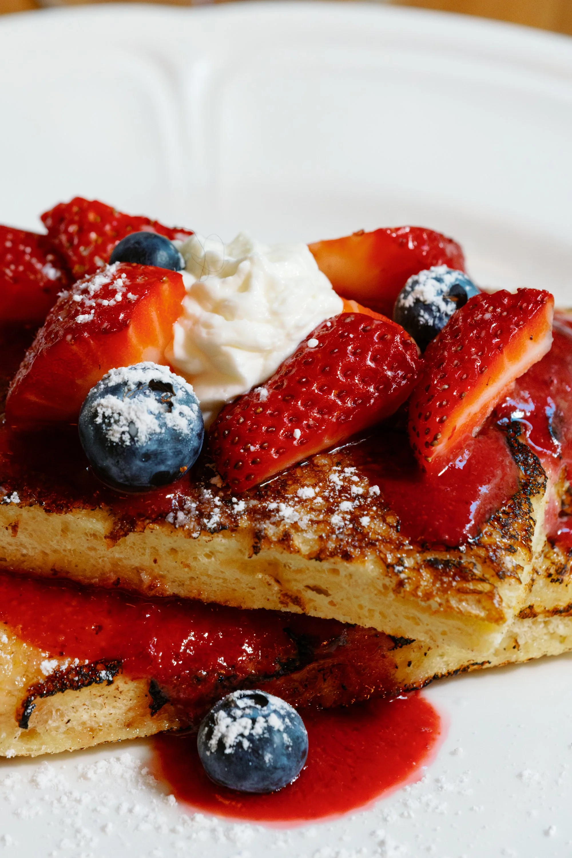 Close-up of a slice of French toast topped with strawberries, blueberries, whipped cream, powdered sugar, and strawberry syrup on a white plate.
