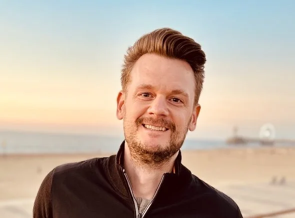 A smiling man with short brown hair and a beard, wearing a black jacket, standing on a beach at sunset.
