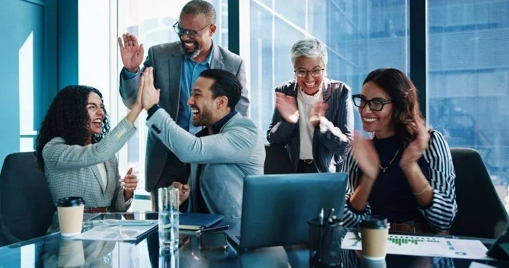 A group of five business professionals celebrating and giving high-fives in a modern office setting, with a large window and city view in the background.