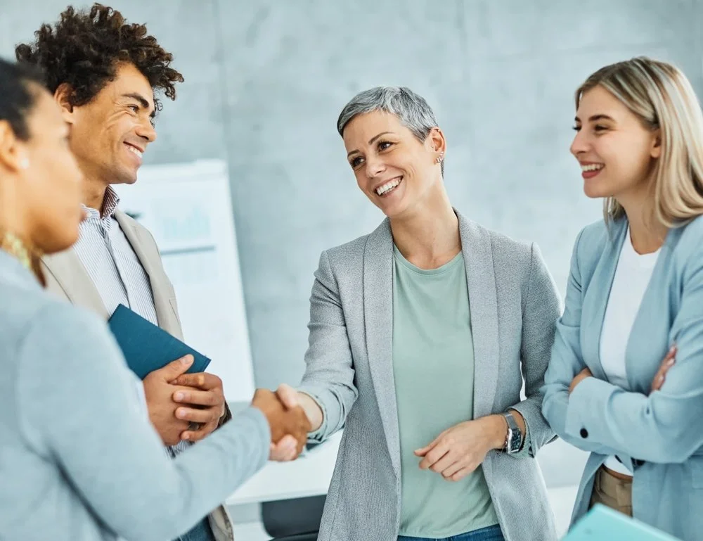 Four professional women and one man engaging in a business handshake in an office setting, smiling and dressed in business casual attire.