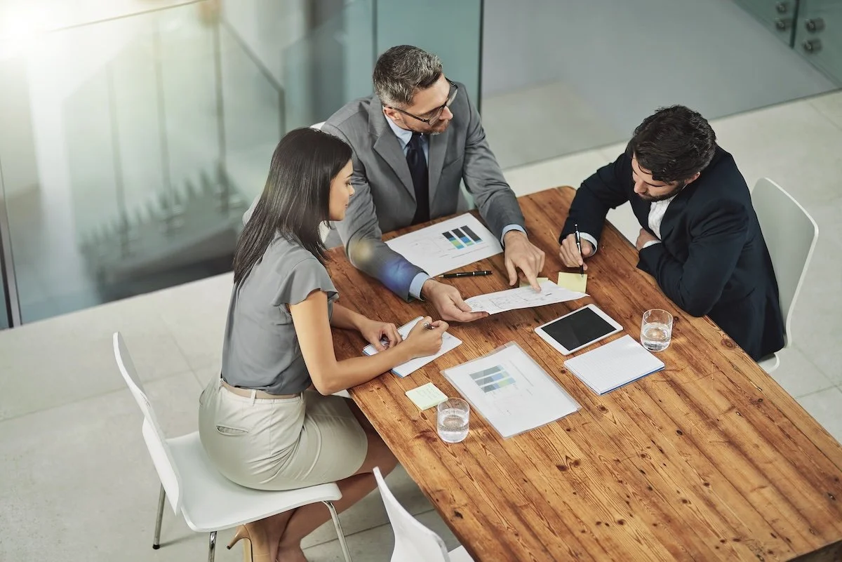 Three business professionals, engaged in a meeting around a wooden table reviewing documents and notes on healthcare organization's operational success.
