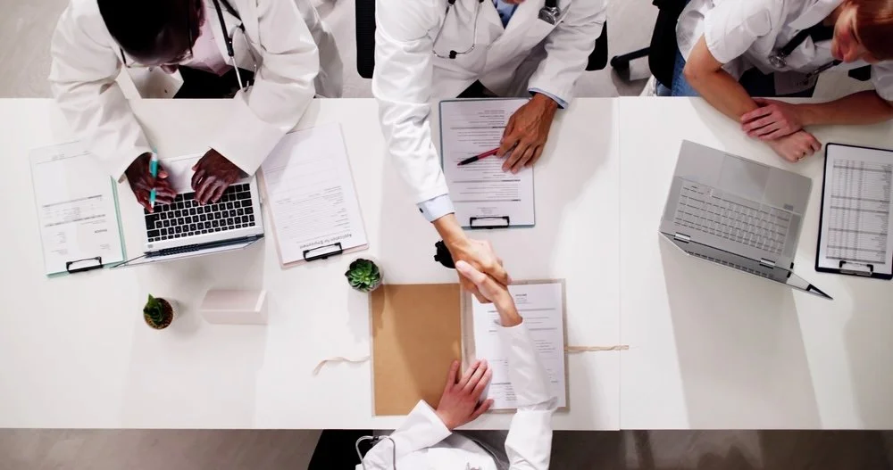 Top-down view of medical professionals in white coats shaking hands over a conference table with documents, laptops, working towards maximizing outcomes for patients..