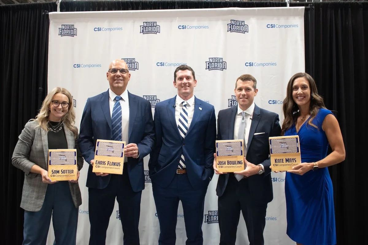 Five professionally dressed individuals standing in front of a white backdrop with the logos of North Florida and CSI Companies, smiling and holding plaques.