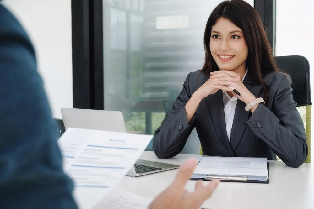 A young woman dressed in business attire smiling and sitting at a desk during a interview in an office.