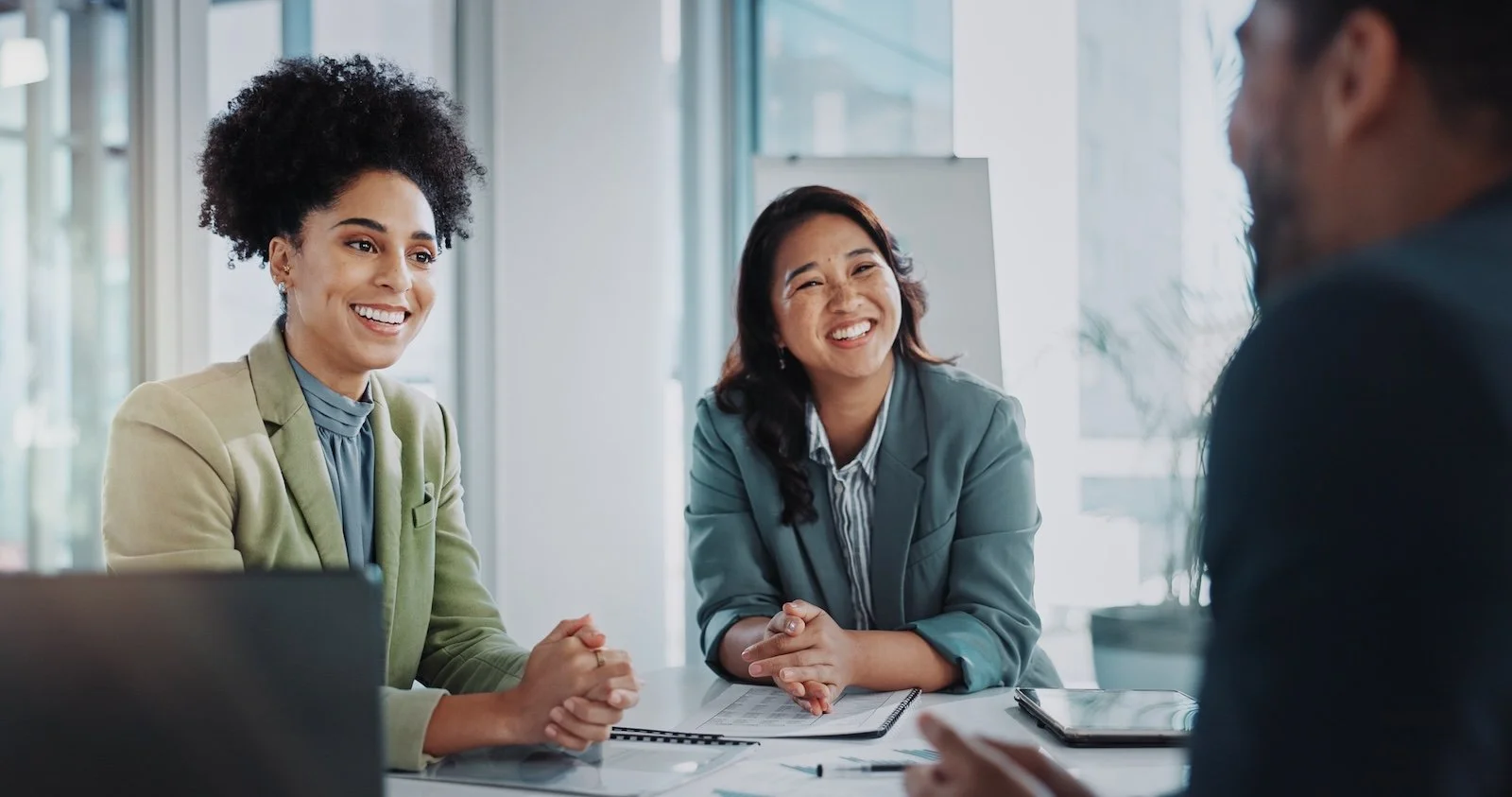 Two business professional women smiling and talking to a man about career growth in a meeting room with documents and laptops.
