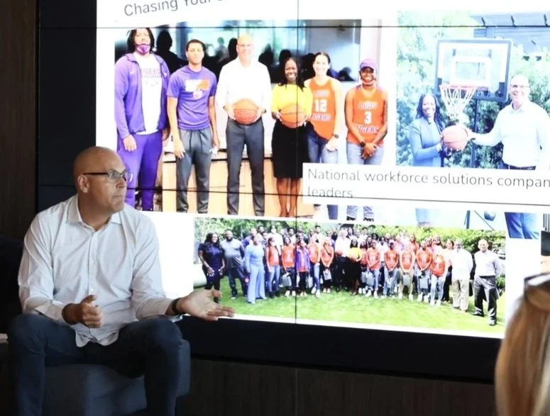 CEO of CSI, Chris Flakus sitting and speaking in front of a large screen displaying group photos of community service, at a presentation .