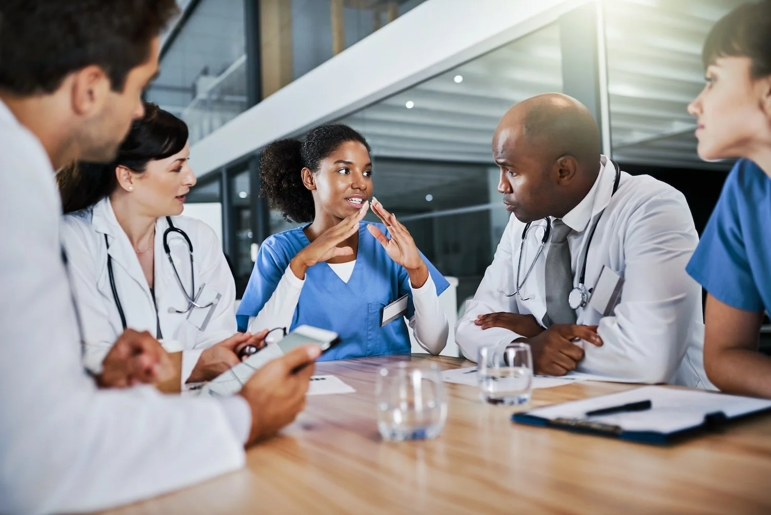 A diverse group of five healthcare professionals, including three women and two men, are engaged in a discussion at a medical meeting. They are sitting around a wooden table with glasses of water, notebooks, and a pen, inside a modern medical facility.