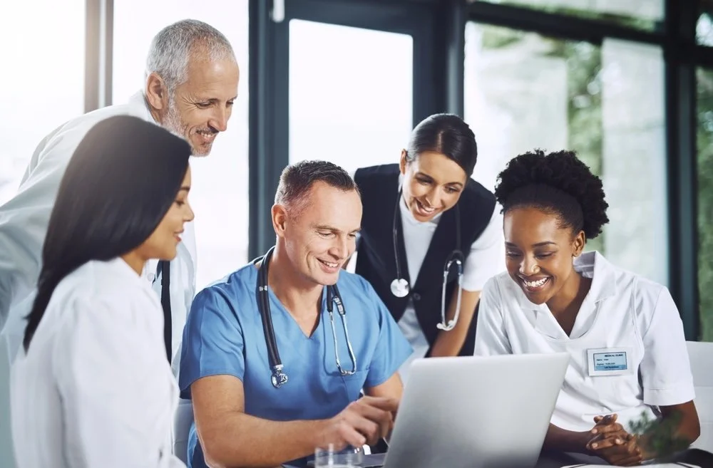 Group of medical professionals, gathered around a laptop discussing operation change management. They are smiling and engaged in a team meeting.