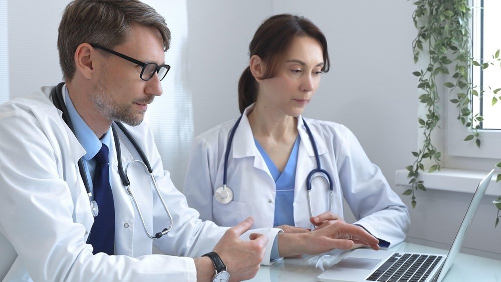 Two doctors, a man and a woman, in white coats with stethoscopes, looking clinical data reviewing for data accuracy together in a medical office.