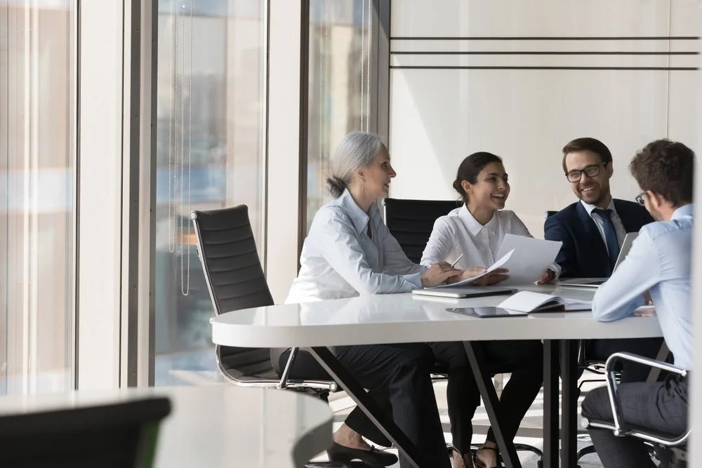 Four healthcare IT business professionals are sitting at a modern conference table having a discussion about technical services in a bright office with large windows.