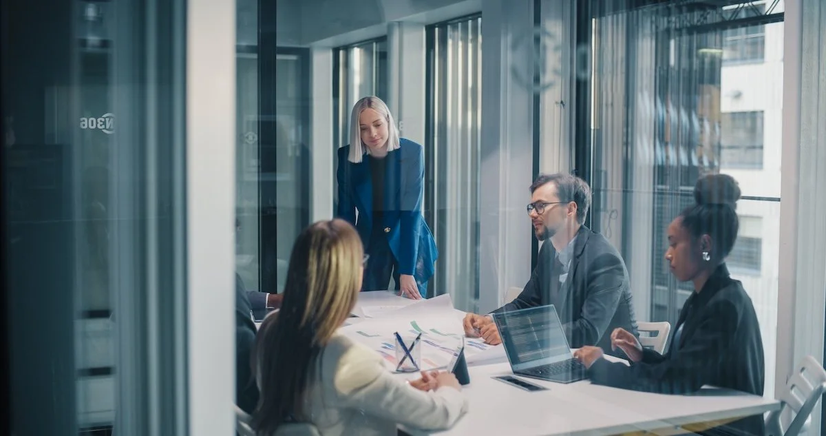 Five healthcare IT team members at a business meeting in a modern conference room with large windows, one woman standing and four seated, discussing EHR integration documents and using laptops.
