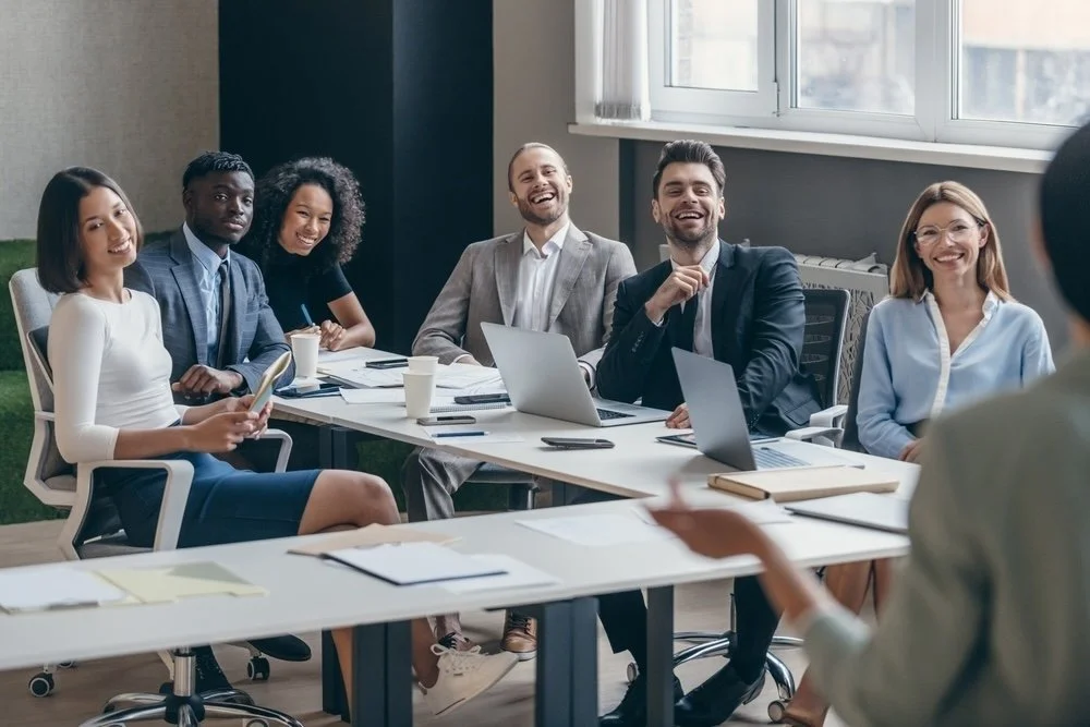 A diverse group of six healthcare IT consultants sitting around a conference table, smiling and laughing during a meeting in a modern office with large windows discussing Epic implementation support.