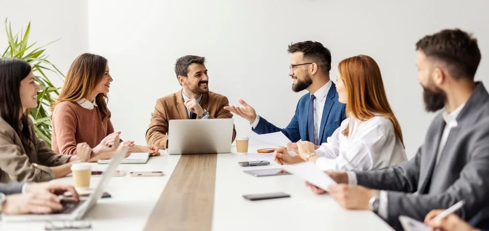 Six diverse healthcare IT team members, engaged in a discussion about epic community connect expansion in a modern conference room with a large wooden table, laptops, and tablets,