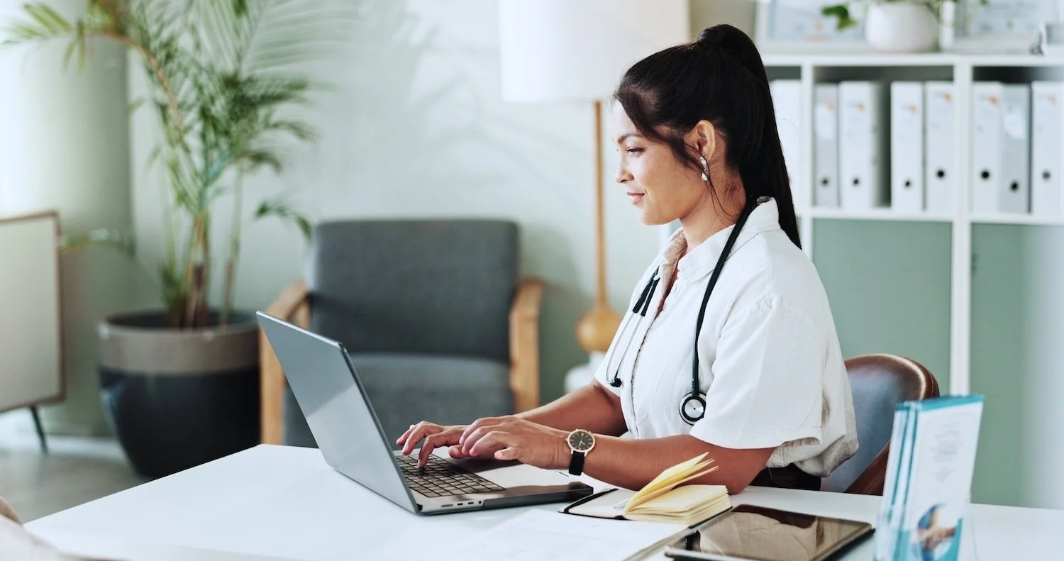 A female doctor sitting at a desk, working confidently on her laptop with a stethoscope around her neck.