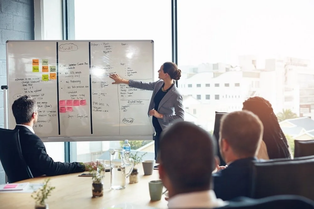 Team of healthcare IT leaders meeting to look at data and community connect methodology on a whiteboard, in a bright meeting room with tall windows. 