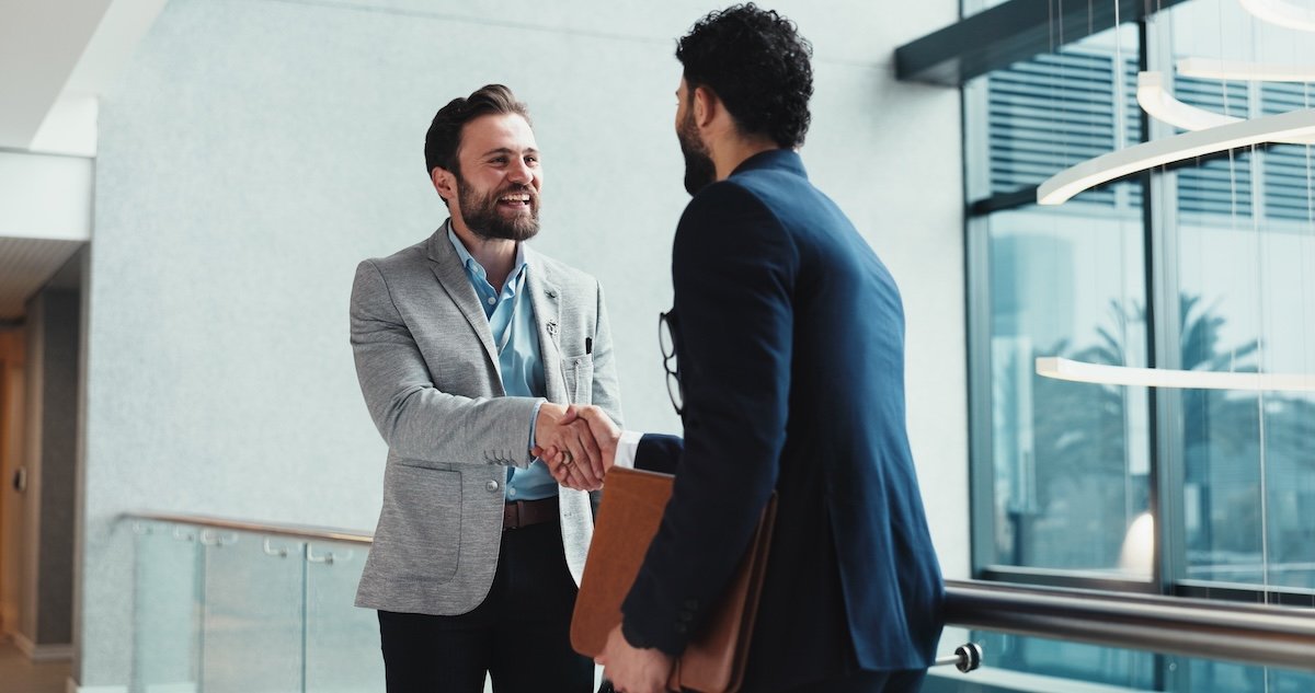 Two men in business attire shake hands in a modern office building, preparing for interview.