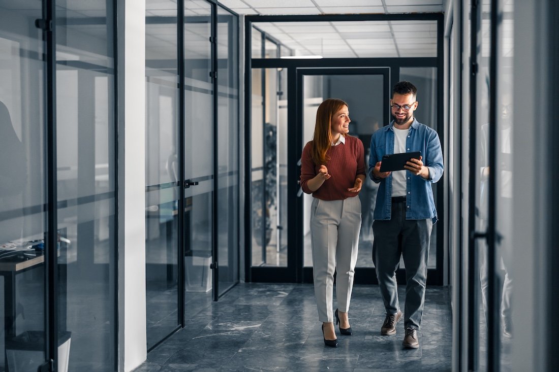Two business professionals, walking and smiling in a modern office hallway with glass walls, looking at a tablet.