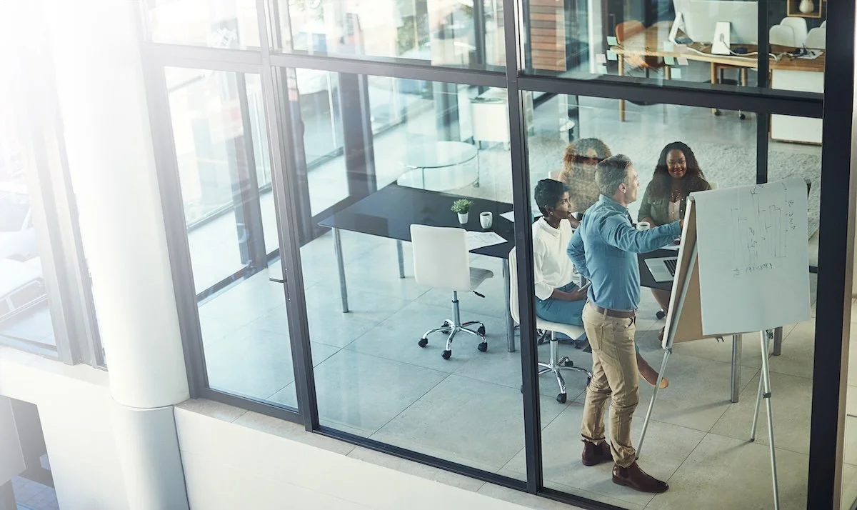 A group of healthcare IT leaders in a modern office meeting room with glass walls, with a man presenting at a whiteboard while other team members listen and look on.