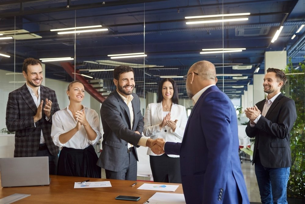 A business meeting with six professionals in an office, with two men shaking hands.