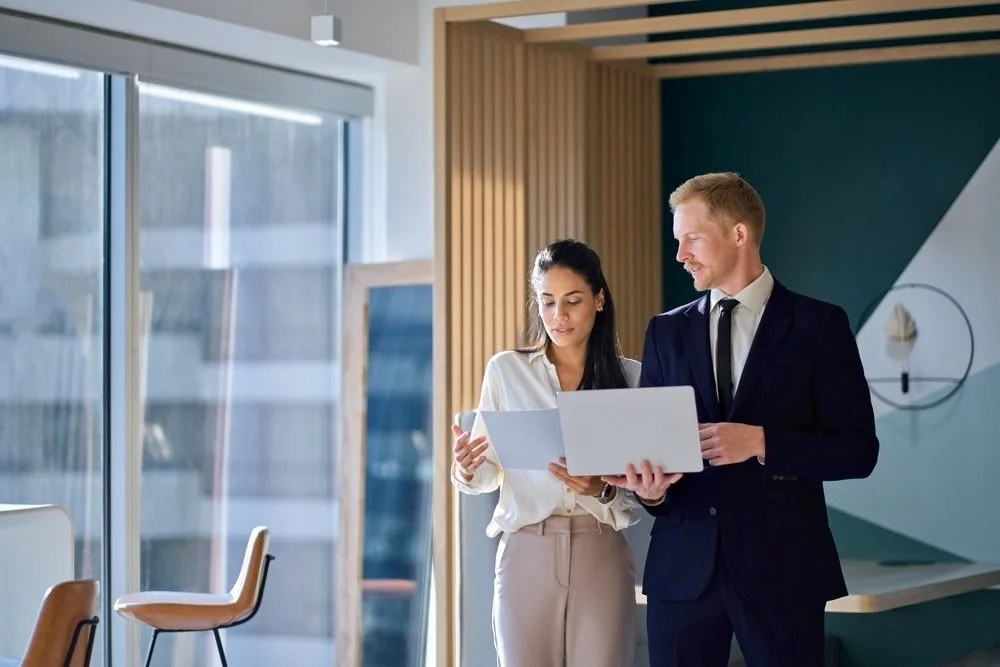 Two business professionals, a woman and a man, standing in a modern office, looking at documents and a laptop together.