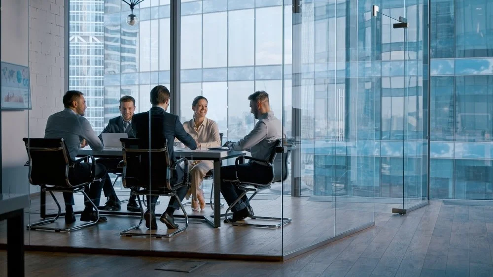 Business team in a glass-enclosed conference room engaged in discussion, with modern city skyline visible through large windows.