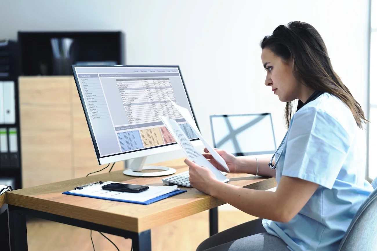 Nurse sitting in a modern office, reviewing medical coding charts on her desktop and auditing information on documents
