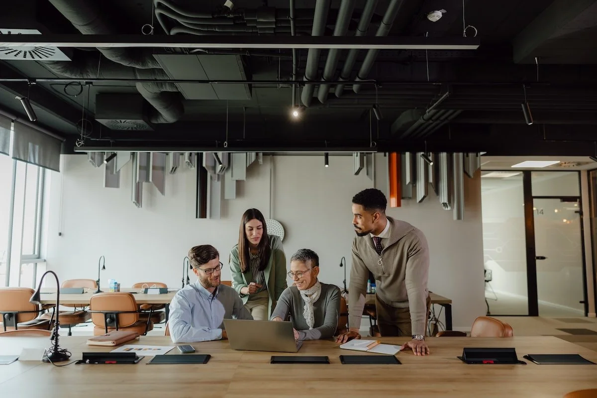 Four corporate professionals in an office collaborating around a laptop at a large conference table.