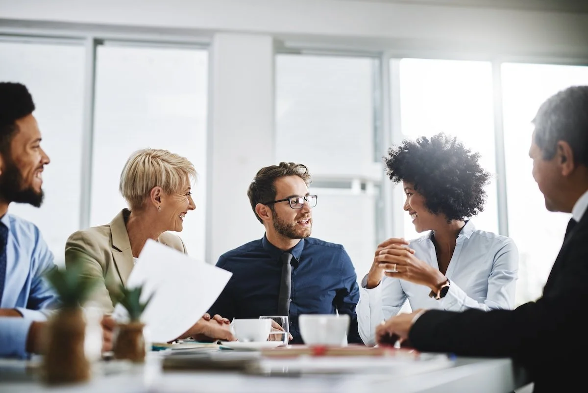 A diverse group of five corporate team members in a bright office, engaged in friendly conversation around a table with documents and coffee cups.