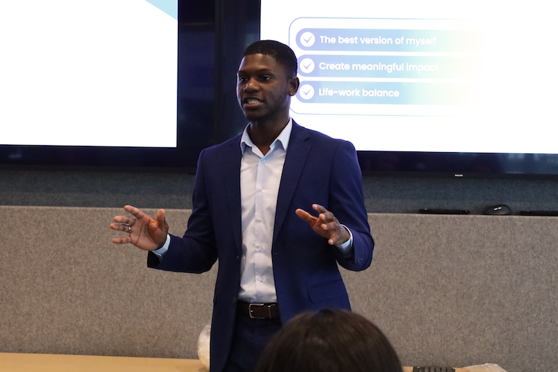 A CSI Connect intern young man, in a blue suit giving a presentation in front of a screen with bullet points about self-improvement and work-life balance.