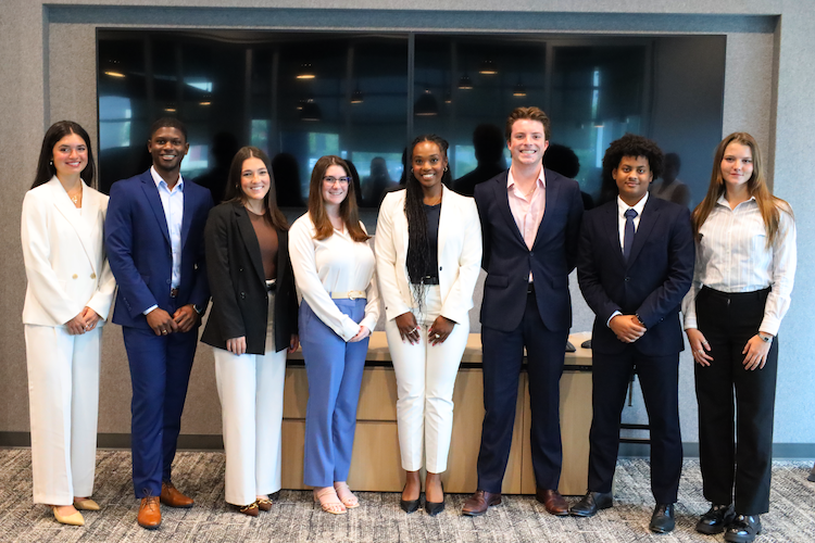 Group of nine CSI connect interns, standing in a row indoors in front of a large glass window, dressed in business attire, smiling at the camera.