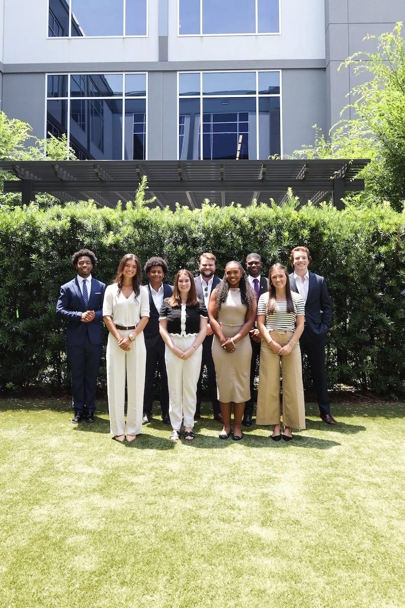 Group of nine CSI Connect interns, standing outdoors in front of a green hedge and a modern glass building, dressed in business casual and formal attire.