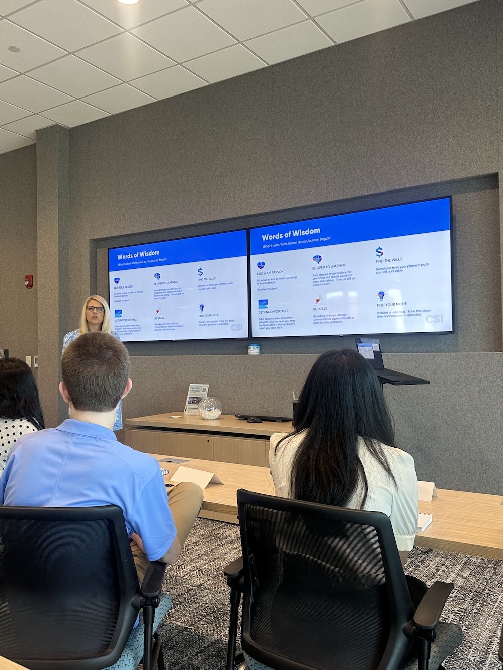 CSI leadership member presenting to a group of interns in a conference room with two large screens displaying a slide titled "Words of Wisdom", discussing about personal growth and soft growth.. 