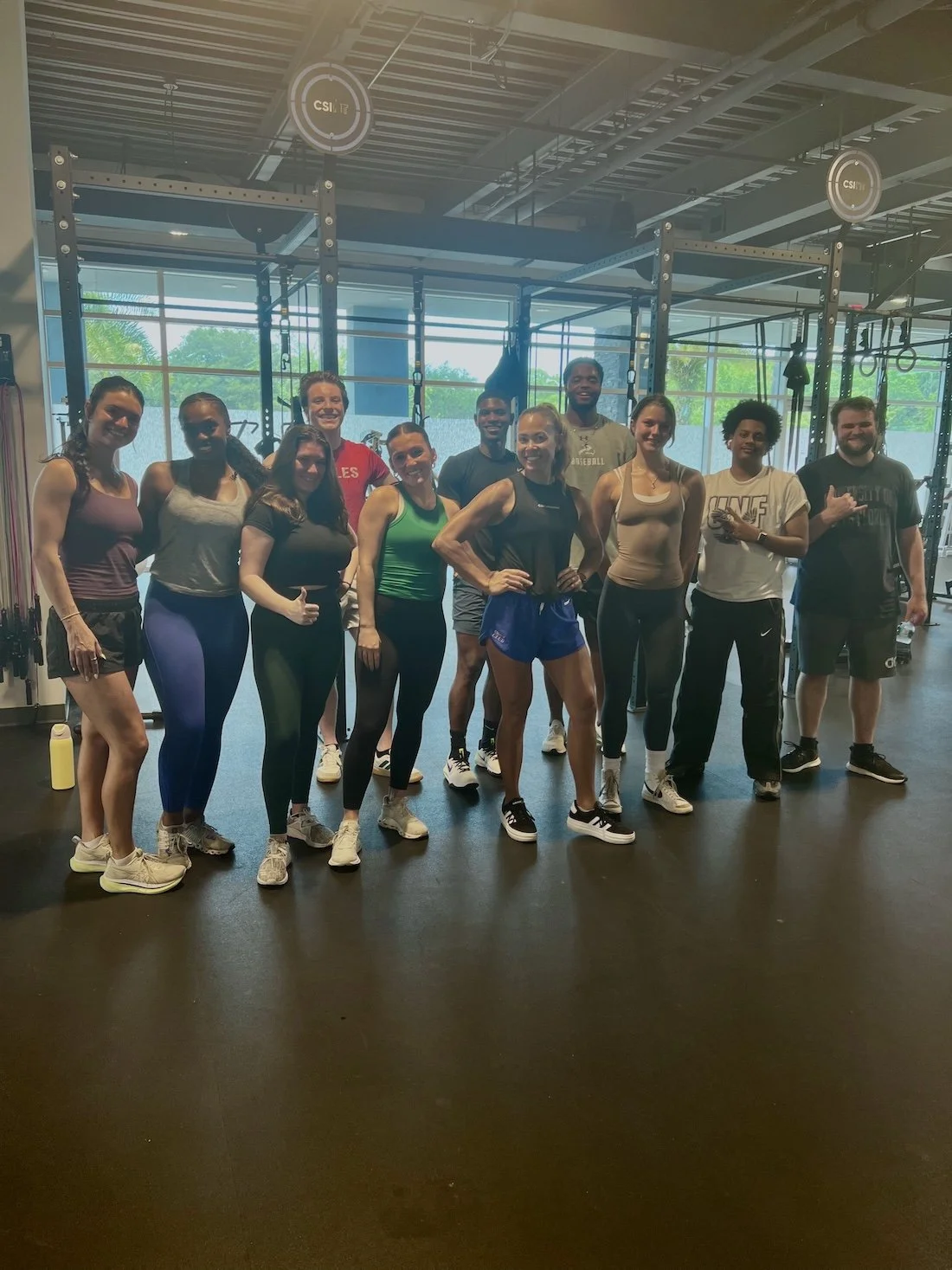 A group of diverse CSI connect interns and a physical trainer standing together inside a gym, smiling post workout, with gym equipment and large windows in the background.