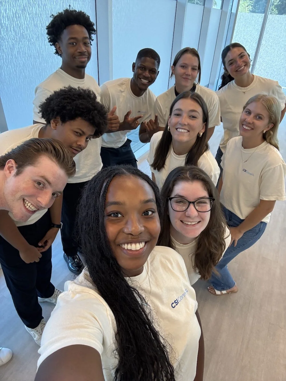 A diverse group of CSI connect interns smiling and posing for a selfie indoors, with glass windows in the background.