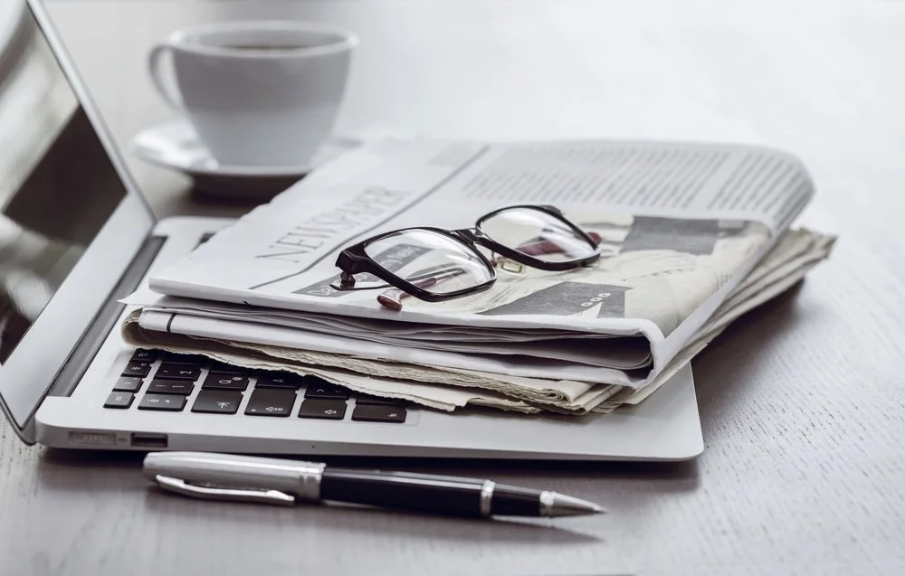 Desk with laptop, newspapers, reading glasses, pen, and a coffee cup.