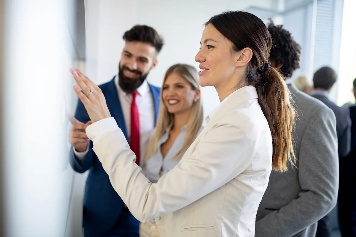 A corporate team memeber in a white blazer is writing on a whiteboard during a meeting, with three colleagues observing and smiling.