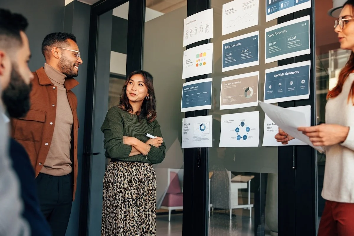 Four diverse corporate team members having a discussion in front of a presentation board with charts and graphs on business forecasts and strategies.