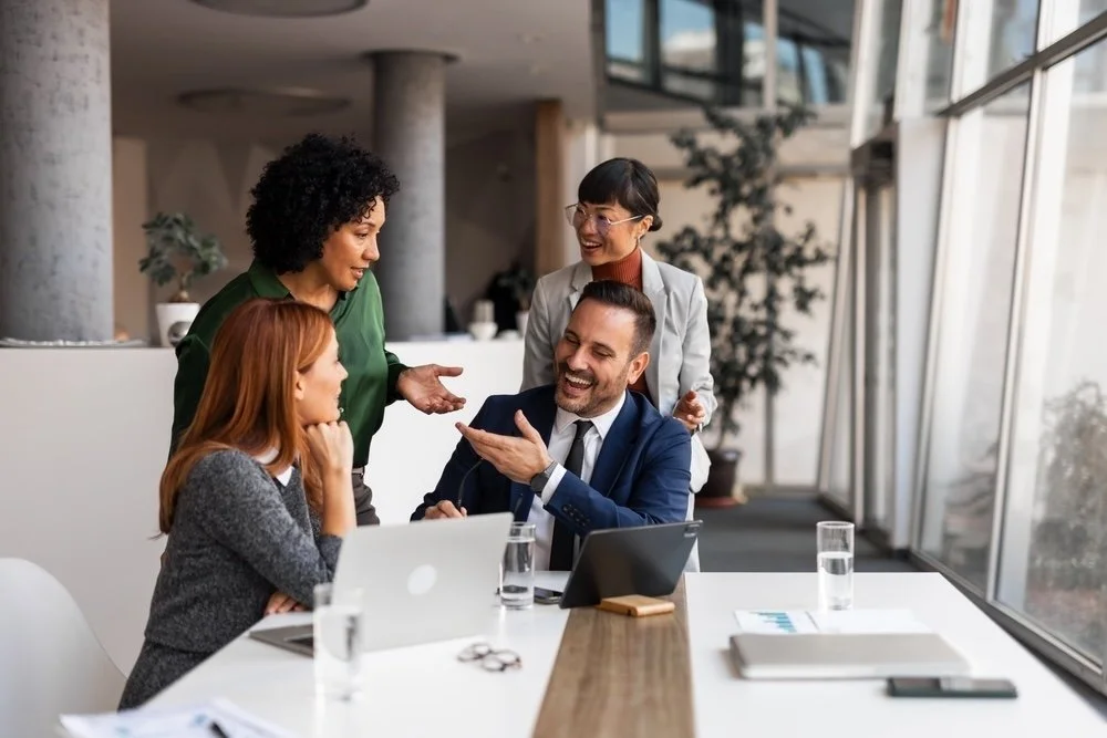Four diverse business professionals in a modern office, engaged in a friendly discussion, with laptops, tablets, and documents on the table.