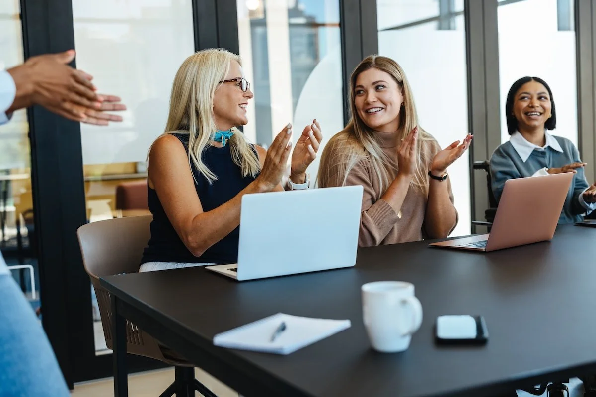 Three business professional women in a meeting room clapping and smiling, with laptops.