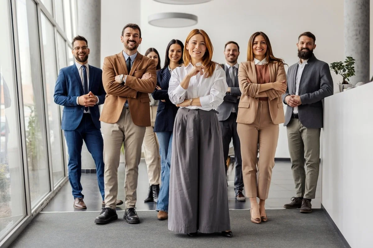 A group of seven diverse professionals standing confidently in a modern office space, smiling at the camera.