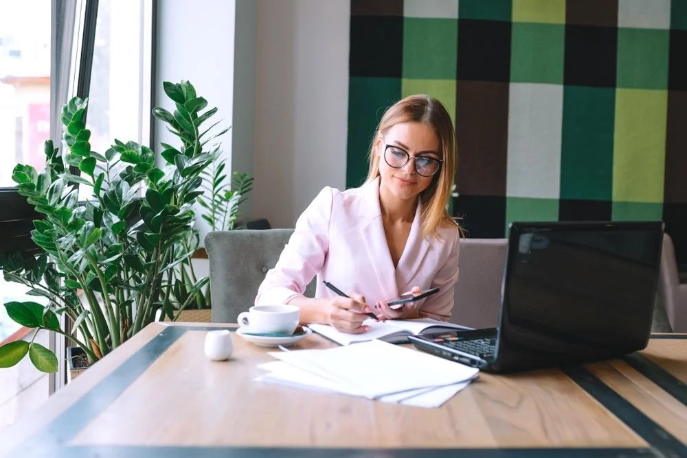 A woman healthcare IT consultant sitting at a wooden table in a bright room, working on a laptop, holding a smart phone