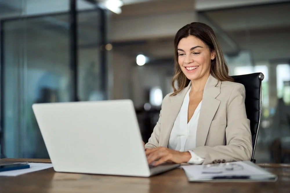 A woman in business attire sitting at a desk, smiling while working on her laptop in an office.