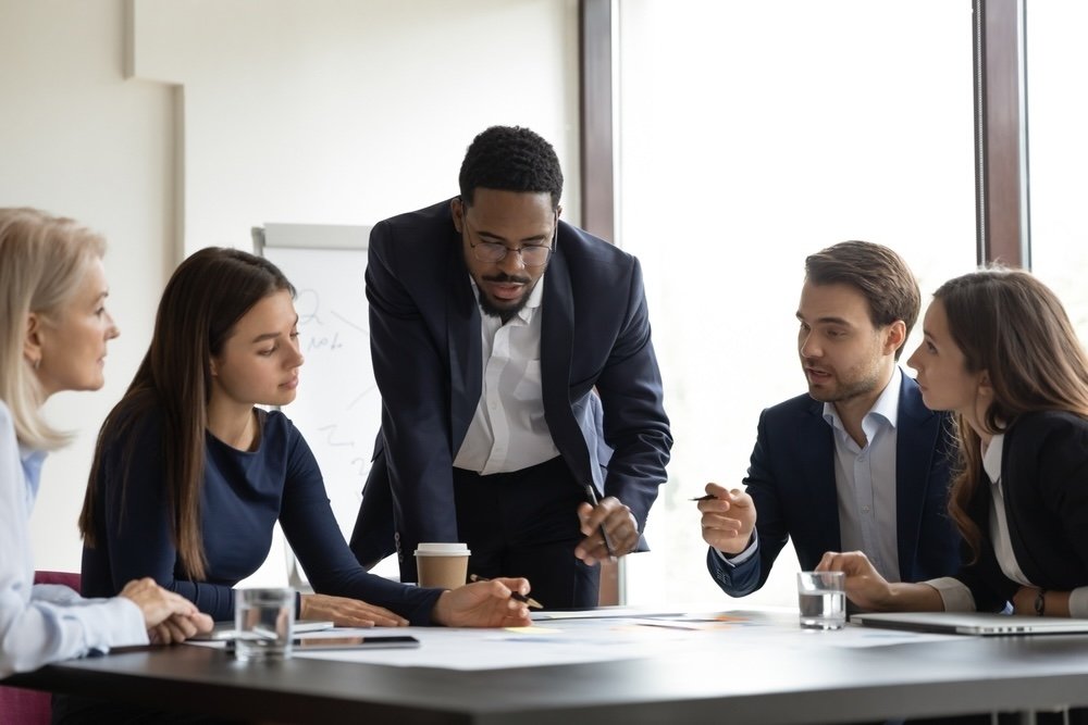 Business meeting with six healthcare IT team members around a table, one man in a suit standing and talking to the others about supporting post go-live challenges.