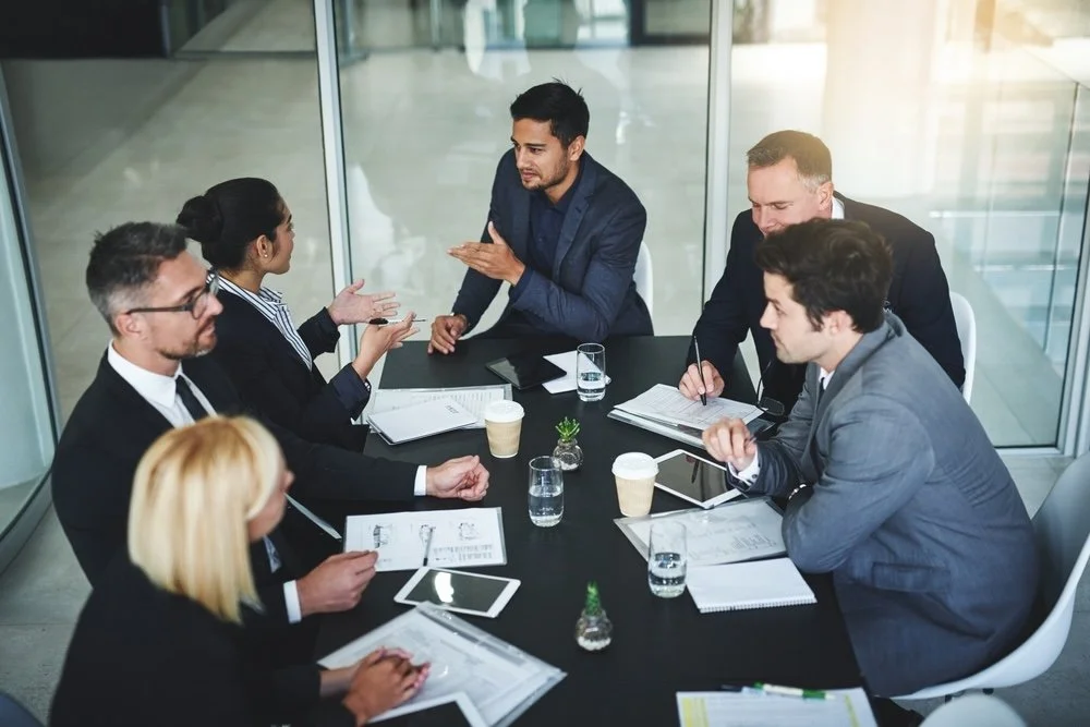 Business meeting with six people seated around a black conference table having a discussion, with documents, tablets, notebooks, and coffee cups on the table.