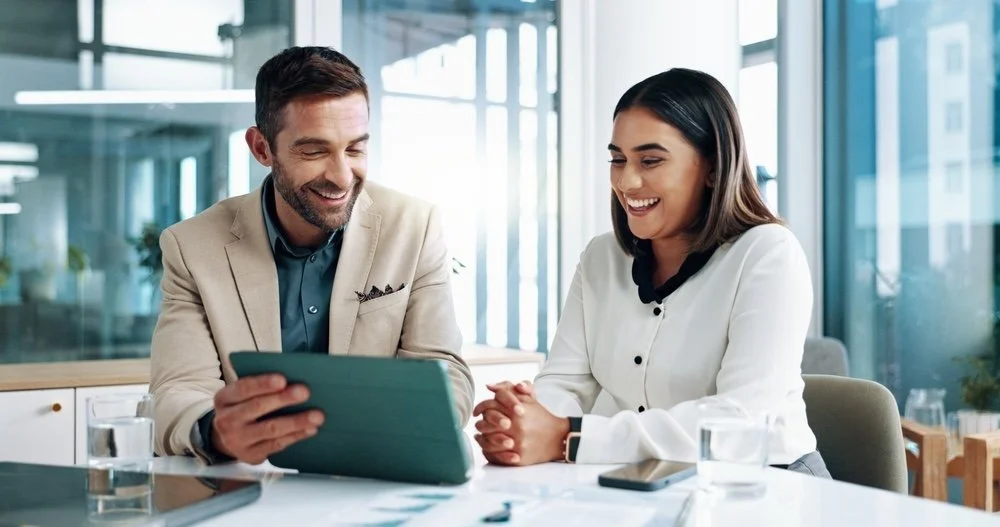 Two healthcare IT consultants eagerly looking at tablet with training project plan details, white sitting down at a table in a modern office.