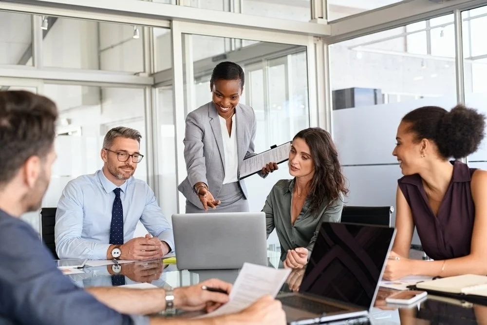 A diverse group of five healthcare IT professionals, meeting in a modern office at a table with laptops and documents reviewing Epic training programs.