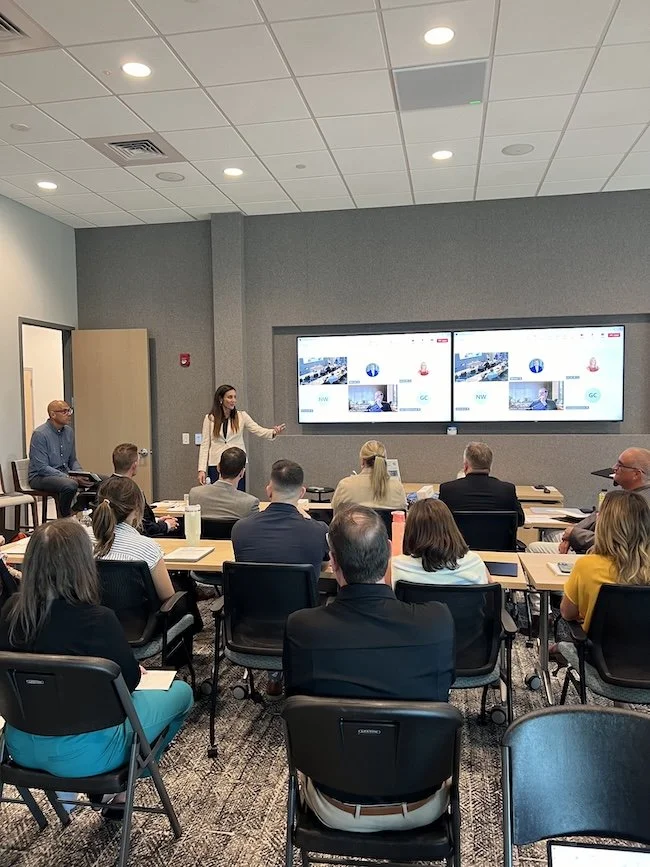 Company wide team meeting inside a modern classroom, with leader speaking to both online and in-classroom team members