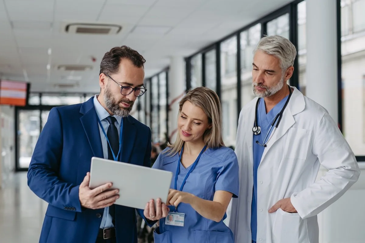 Healthcare consultant reviewing documentation on a tablet with a nurse and a doctor in a hospital hallway.